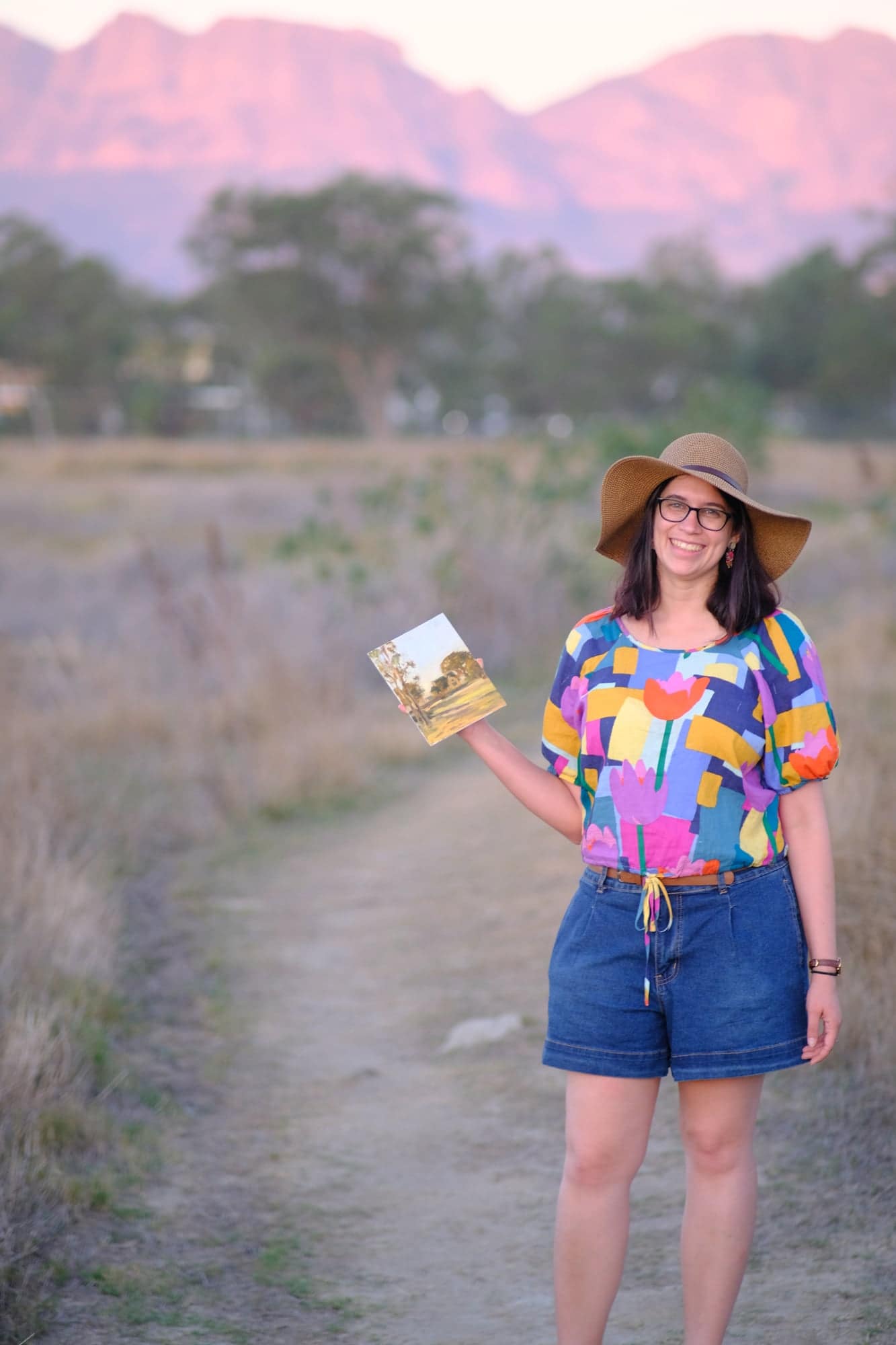 Tiana Goosen holding one of her paintings in a field at sunset, with the mountains behind her